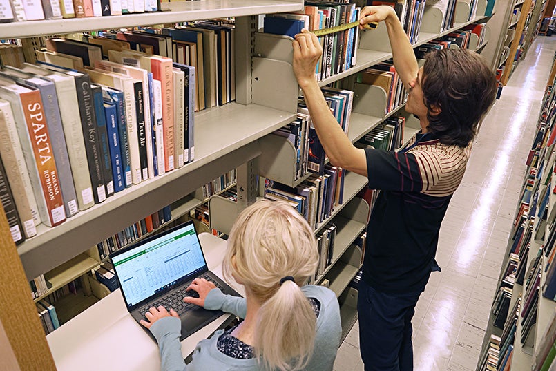 A library staff member measures books in the stacks while another record data on a laptop