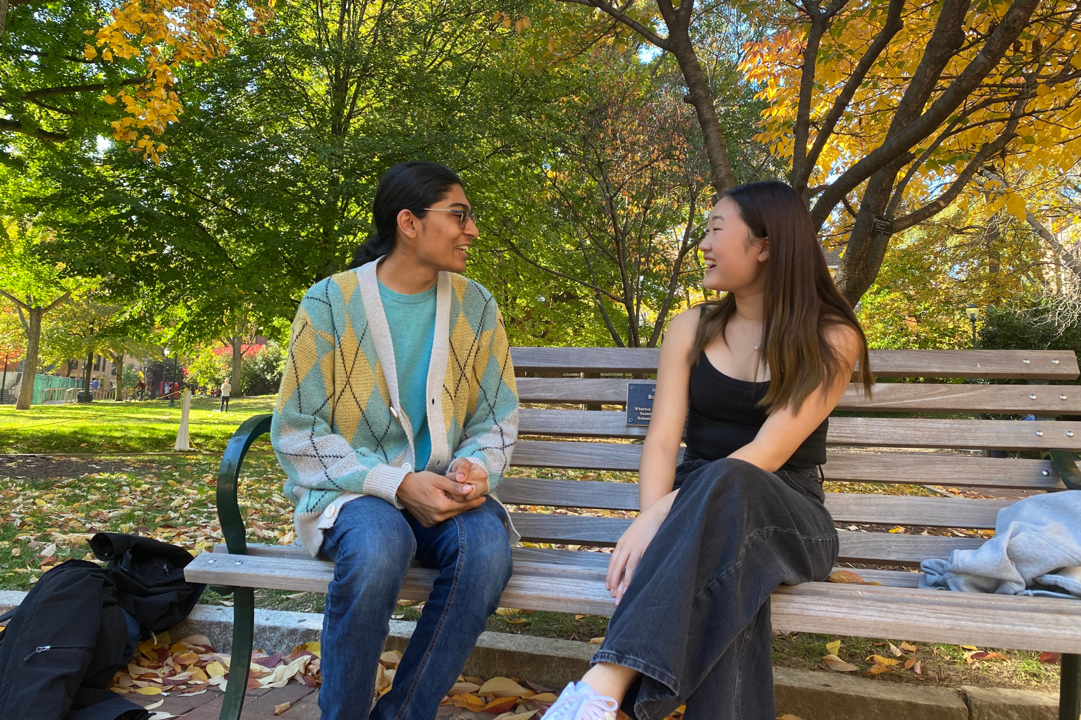 Two students sit on a bench near a tree on a sunny autumn day, talking enthusiastically to each other.