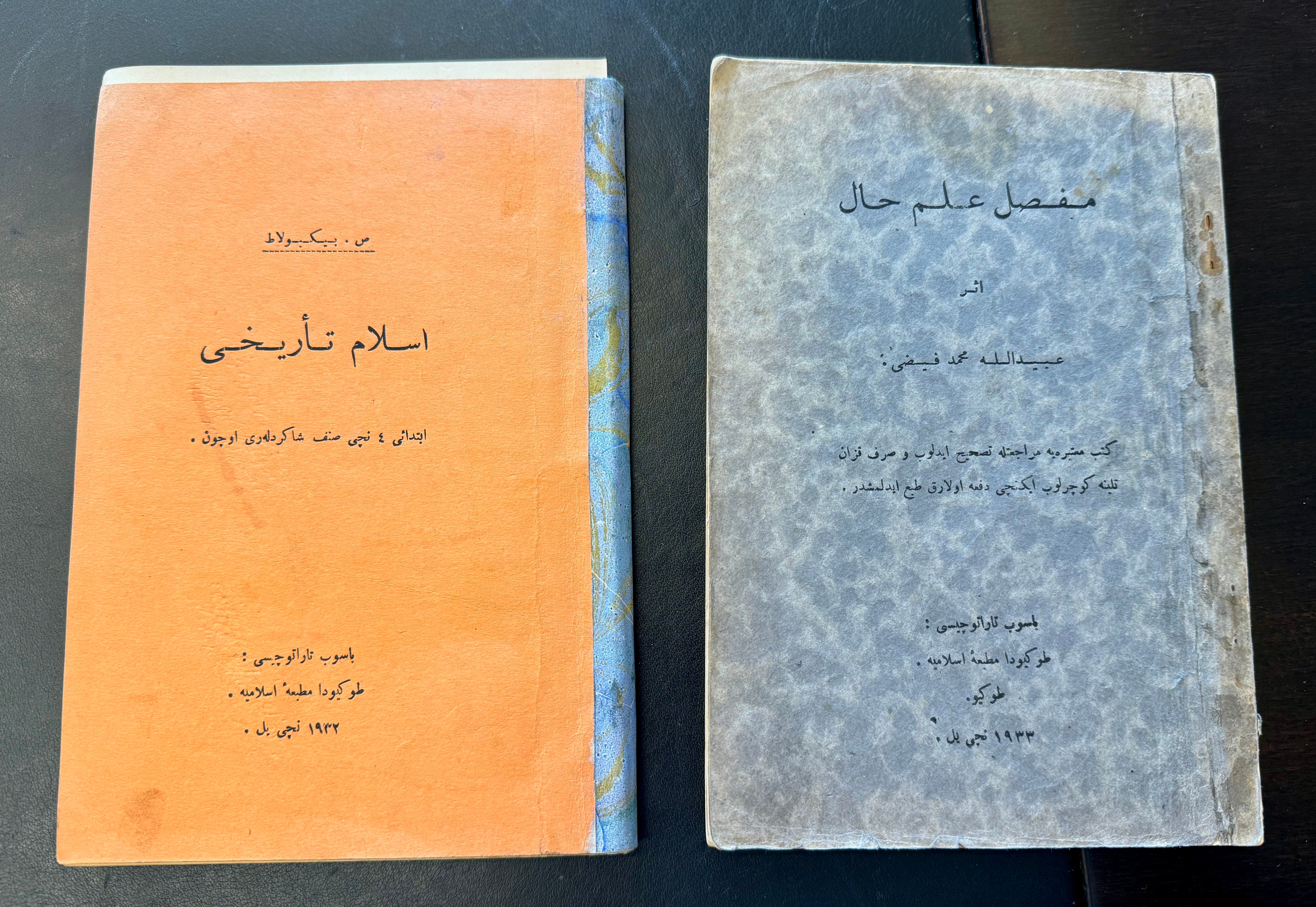 Two books are placed side by side. Both have Arabic text on their covers.  The book on the left has an orange cover with blue binding, while the one on the right has a greyish-blue cover with a textured appearance. 