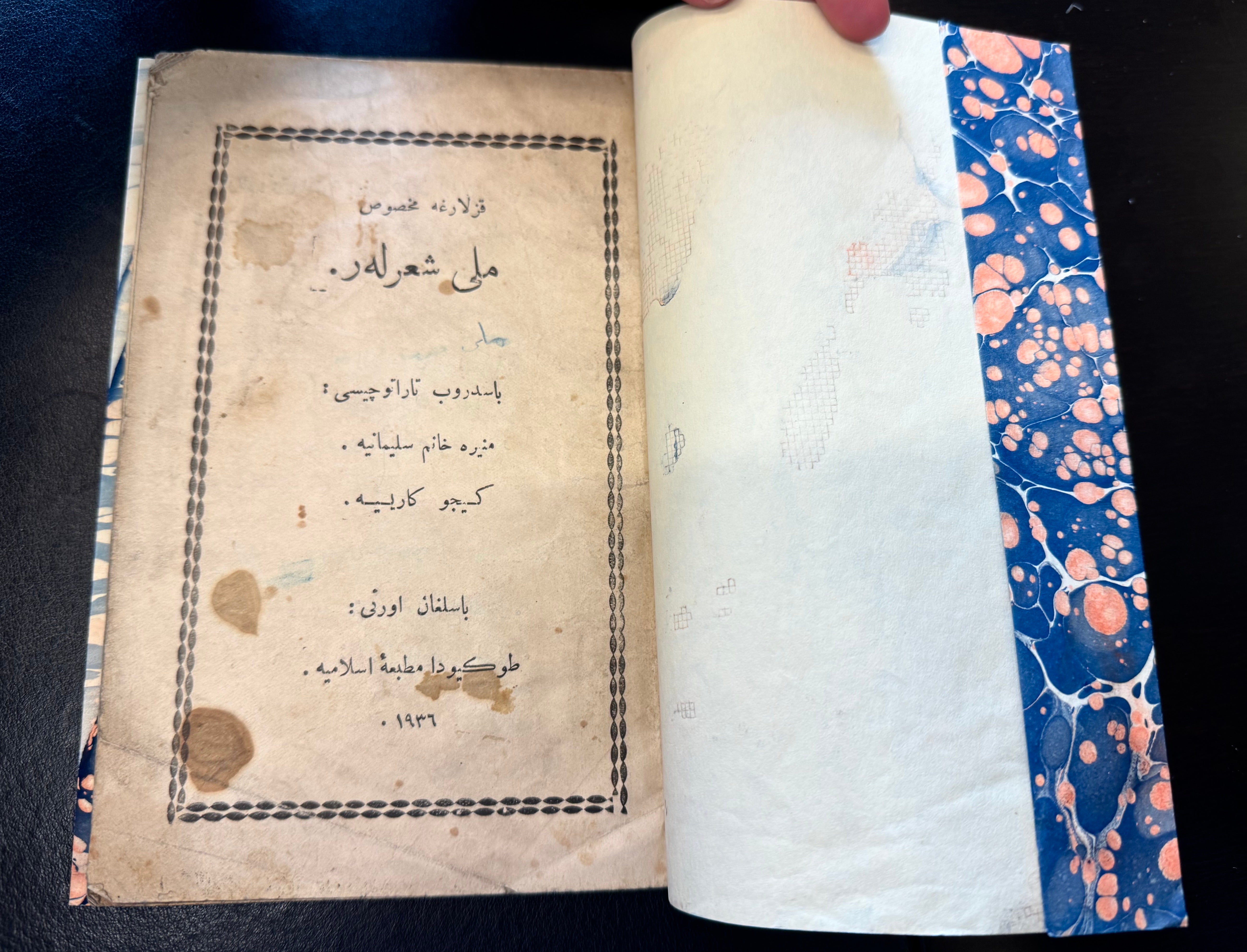A hand is shown turning the page of an old book with Arabic script. The visible page has Arabic text surroundeid by a decorative border, and there are some stains on the page indicating its age. The paper appears aged and slightly yellowed.
